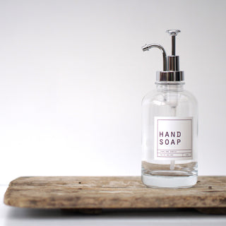 An empty clear glass bottle with a silver pump lid, labeled 'HAND SOAP', placed on a wooden tray.