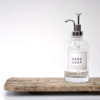 An empty clear glass bottle with a silver pump lid, labeled 'HAND SOAP', placed on a wooden tray.
