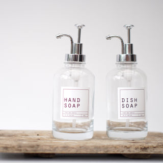 An empty clear glass bottle with a silver pump lid, labeled 'HAND SOAP', placed on a wooden tray.