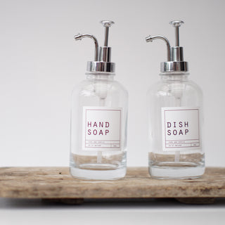 An empty clear glass bottle with a silver pump lid, labeled 'HAND SOAP', placed on a wooden tray.