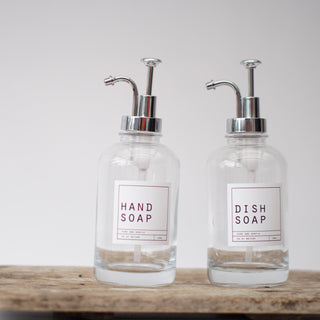An empty clear glass bottle with a silver pump lid, labeled 'HAND SOAP', placed on a wooden tray.