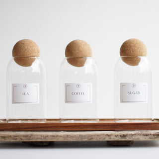 Three clear glass jars with cork ball lids, labeled 'COFFEE', 'SUGAR', and 'TEA', placed on a wooden tray.