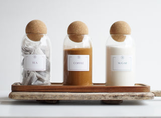 Three clear glass jars with cork ball lids, labeled 'COFFEE', 'SUGAR', and 'TEA', placed on a wooden tray.