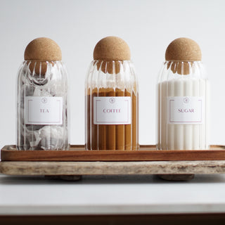 Three clear glass jars with cork ball lids, labeled tea, coffee, and sugar displayed on a wooden tray.