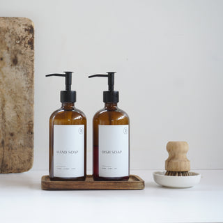 Two brown bottles with pump dispensers on a wooden tray, next to a wooden cutting board and a wooden brush.