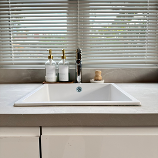 A clear glass bottle set with wooden tray, pot brush, and ceramic dish, displayed on a kitchen counter with blinds and light strip in the background.