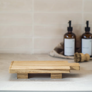 Wooden soap dish on a bathroom counter with bottles and a brush in the background.