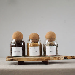 Three clear glass jars with cork ball lids, labeled 'COFFEE', 'SUGAR', and 'TEA', placed on a wooden tray.