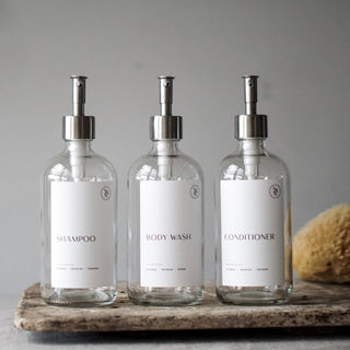 Three clear glass bottles with silver stainless steel dispenser pump lids, labeled SHAMPOO, CONDITIONER, and BODY WASH, displayed on a textured surface.