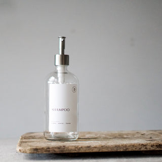 Three clear glass bottles with silver stainless steel dispenser pump lids, labeled SHAMPOO, CONDITIONER, and BODY WASH, displayed on a textured surface.