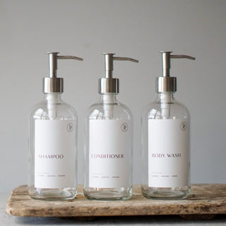 Three clear glass bottles with silver stainless steel dispenser pump lids, labeled SHAMPOO, CONDITIONER, and BODY WASH, displayed on a textured surface.