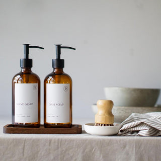 Two amber glass soap dispensers with black pump lids on a wooden tray, accompanied by a white pot brush on a kitchen counter.