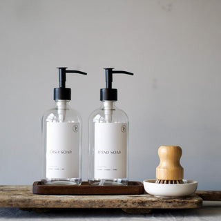 Two clear glass bottles with black pump lids on a wooden tray, accompanied by a wooden pot brush and a ceramic dish, against a grey background.
