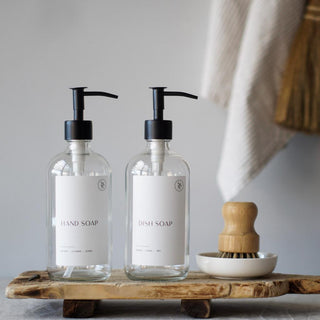 Two clear glass bottles with black pump lids on a wooden tray, accompanied by a wooden pot brush and a ceramic dish, against a grey background.