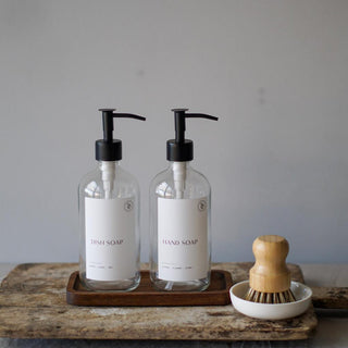 Two clear glass bottles with black pump lids on a wooden tray, accompanied by a wooden pot brush and a ceramic dish, against a grey background.