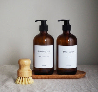 Two amber glass soap bottles with black pump lids labeled 'HAND SOAP' and 'DISH SOAP' on a wooden tray, alongside a wooden dish brush.