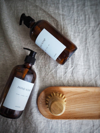Two amber glass soap bottles with black pump lids labeled 'HAND SOAP' and 'DISH SOAP' on a wooden tray, alongside a wooden dish brush.