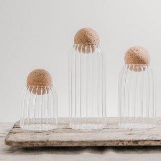 Three clear glass jars with reeded design and cork ball lids on a wooden surface.