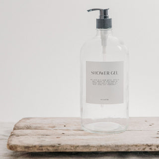 A clear glass bottle with a black pump lid, labeled 'SHOWER GEL', placed on a wooden surface.