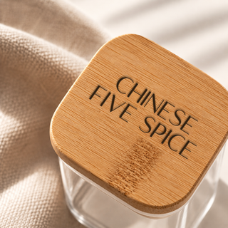 A clear glass spice jar with a square bamboo lid engraved with the words 'CHINESE FIVE SPICE', resting on a textured beige fabric surface.