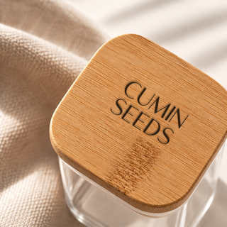 A clear glass jar with a square rounded bamboo lid engraved with 'CUMIN SEEDS', resting on a beige textured fabric under soft natural light.