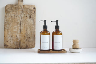 Two amber glass bottles with pump dispensers on a wooden tray, next to a wooden cutting board and brush.
