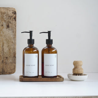 Two brown bottles with pump dispensers on a wooden tray, next to a wooden cutting board and a wooden brush.