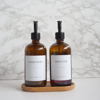 Two amber glass soap dispensers labeled 'Hand Soap' and 'Dish Soap' on a wooden tray with a marble background.