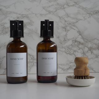 Two brown bottles labeled 'Hand Soap' and 'Dish Soap' with a wooden brush in a white holder on a marble background.