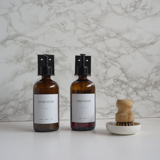 Two bottles of hand soap and dish soap with a wooden brush on a marble background