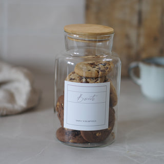 A clear glass cookie jar with a light wood lid, filled with assorted biscuits, on a light marble countertop with a soft-focus kitchen background.