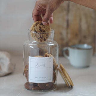 A hand placing a chocolate chip cookie into a clear glass jar labeled 'Biscuits' by Smarty Scramble, with additional cookies inside and a mug blurred in the background.