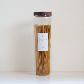 A tall clear glass jar filled with spaghetti, featuring a white label with the brand name 'Sapori di Milano' and a dark brown wooden lid, set against a plain off-white background.