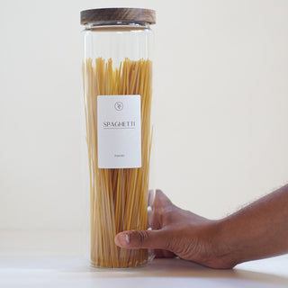 A clear cylindrical glass jar filled with dry spaghetti, topped with a dark wood lid and labeled 'SPAGHETTI,' with a person's hand holding the base against a plain light background.