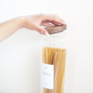A hand with light pink manicured nails reaches down to place a dark wood lid on a clear glass jar filled with spaghetti. The jar has a white label with the word 'SPAGHETTI' printed on it.