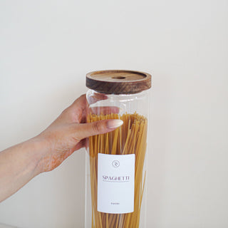 A hand holding a clear glass jar filled with spaghetti, featuring a dark wood lid and a white label that reads 'SPAGHETTI'.