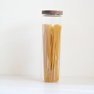 A tall clear glass jar with a dark wood lid, filled with dry spaghetti, set against a plain white background.