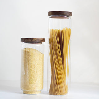 Two clear glass canisters with dark brown wooden lids, one filled with yellow pasta and the other with spaghetti, set against a plain white background.