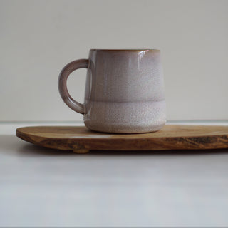 A handmade ceramic mug with a speckled, textured finish in a muted dusty rose and beige color, resting on a rustic wooden board against a neutral light gray background.