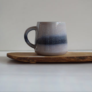 A handmade ceramic mug with a speckled gray and dark blue gradient glaze, resting on a rustic wooden board against a neutral light gray background.