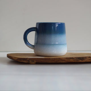 A ceramic mug with a blue-to-white gradient glaze, resting on a rustic wooden board against a neutral gray background.