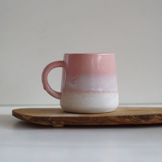 A handmade ceramic mug with a pink and white gradient glaze, resting on a rustic wooden board against a neutral gray background.