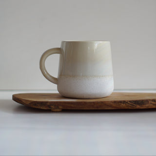 A ceramic mug with a smooth cream-to-white gradient glaze, resting on a rustic wooden serving board against a neutral light gray background.