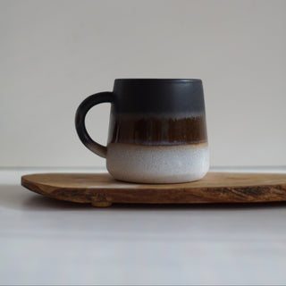 A ceramic mug with a dark brown and black glaze on the upper half and a speckled off-white base, resting on a wooden board against a neutral light gray background.