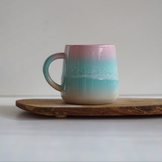 A ceramic mug with a pink-to-turquoise gradient and white speckled wave-like pattern, sitting on a rustic wooden board against a neutral gray background.
