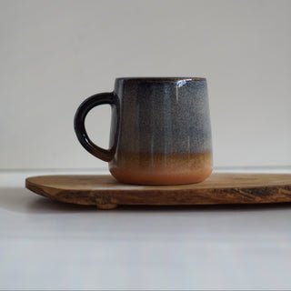 A handmade ceramic mug with a dark gray to warm orange-brown gradient glaze, resting on a rustic wooden board against a neutral light gray background.