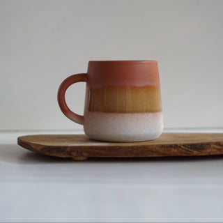 A ceramic mug with a terracotta top, golden-brown middle band, and white speckled base, sitting on a wooden board against a neutral gray background.