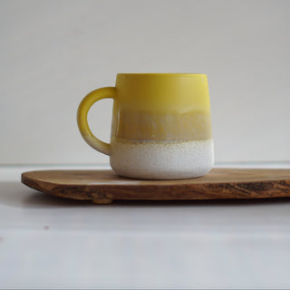 A handmade ceramic mug with a yellow and white gradient glaze, resting on a rustic wooden slice coaster against a neutral gray background.