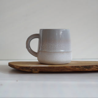 A handmade ceramic mug with a gray to white gradient glaze, resting on a rustic wooden board against a neutral light gray background.