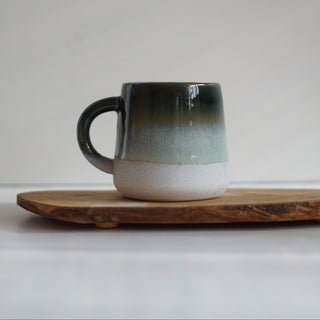 A ceramic mug with a dark green to off-white gradient glaze, resting on a rustic wooden board against a neutral light gray background.